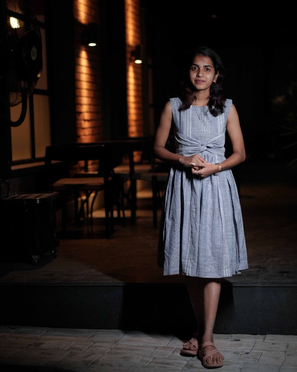 A woman in a beautifully embroidered blue dress stands calmly in a cozy café at night, surrounded by warm ambient lighting.
