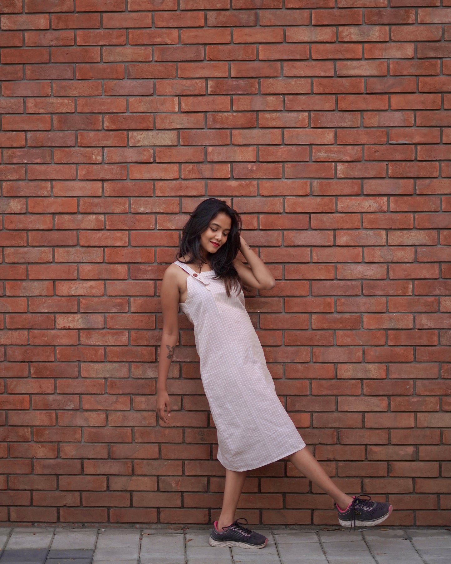 A woman waves her leg playfully while leaning on a brown brick wall, wearing a Lacey Pearl Pinafore with a vibrant pink-black contrast shoe.
