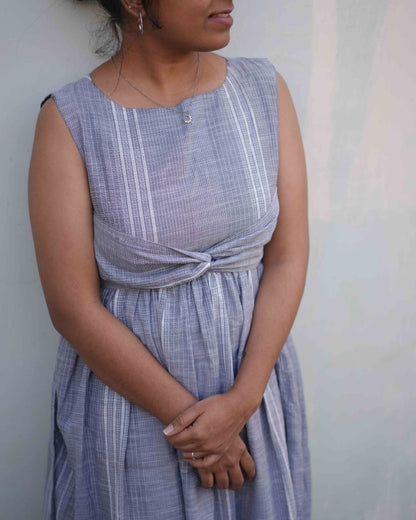 A woman in a comfortable blue dress with an elegant embroidery design from the Vestures collection stands gracefully, gazing into the distance.