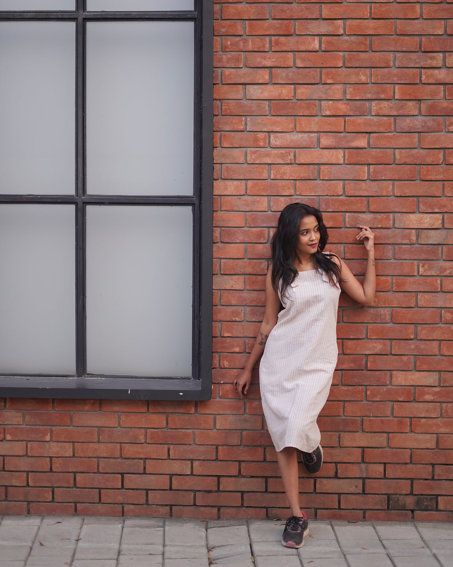 A woman leans on a brown brick wall wearing a Lacey Pearl Color Cozy One-Piece Pinafore, with a vibrant mirror contrast highlighting the dress’s elegance.