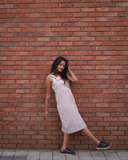 A woman waves her leg playfully while leaning on a brown brick wall, wearing a Lacey Pearl Pinafore with a vibrant pink-black contrast shoe.