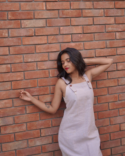 A woman leans over a wall, resting one hand behind her head and the other on the wall, exuding relaxed vibes in a soft linen cotton pinafore.