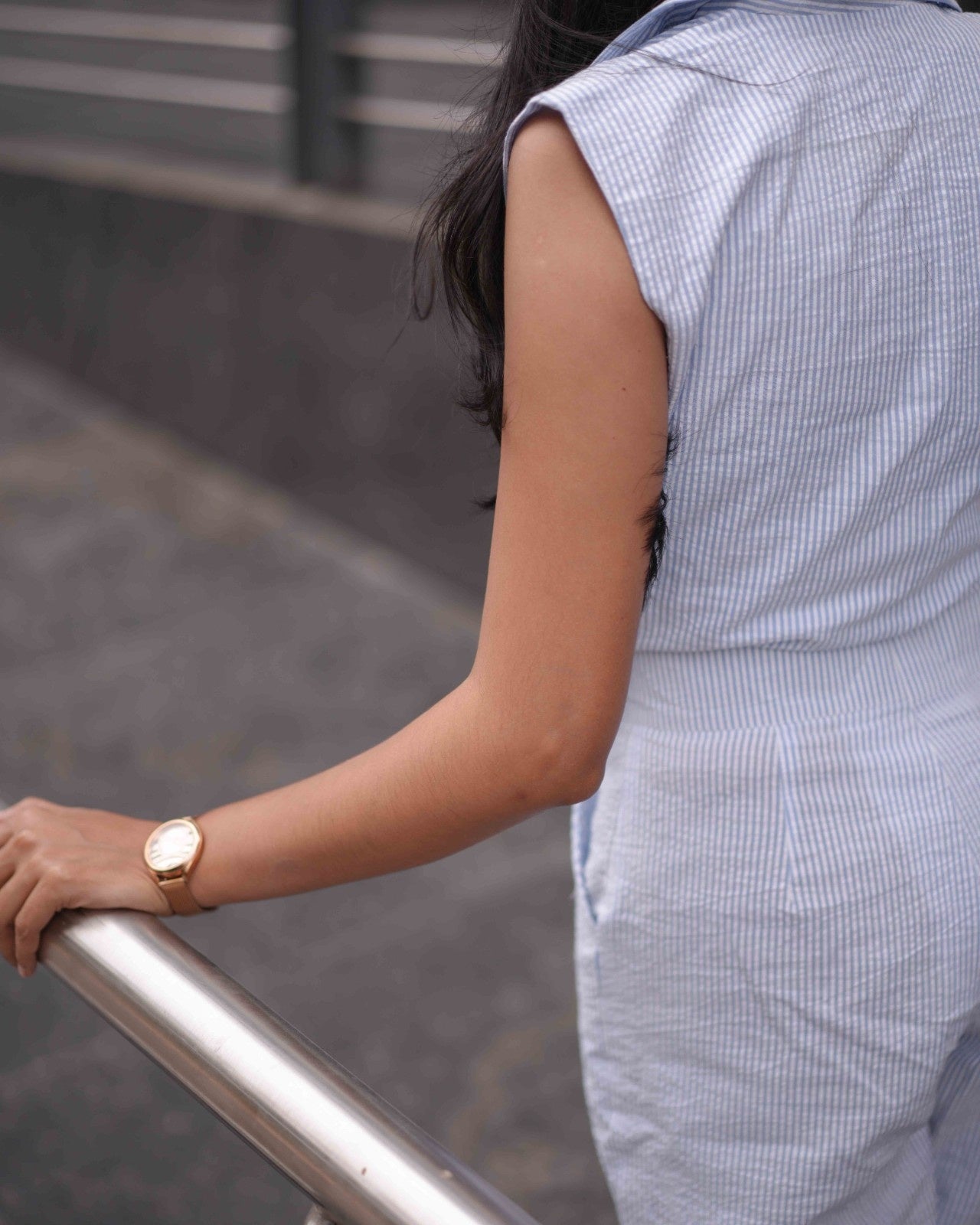 Back left vertical shot of a woman in a striped White and Breeze Blue Jumpsuit, walking up the lawn with a relaxed vibe.