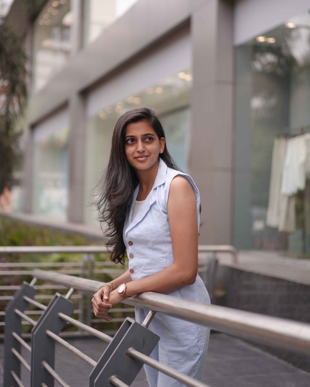Full-body side shot of a woman leaning over iron railings in a White and Breeze Blue Crushed Cotton Jumpsuit, glancing to the left.