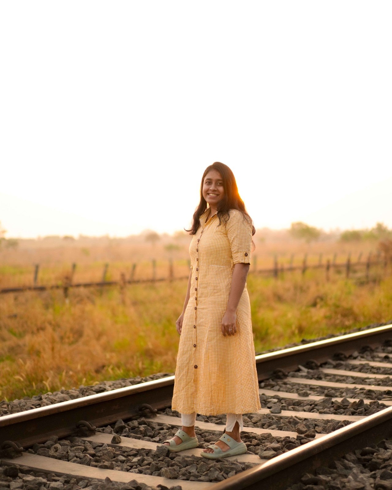 Woman in a Yellow and White Checked Linen Shirt Dress standing on a railway track at dawn, glowing in sunlight.