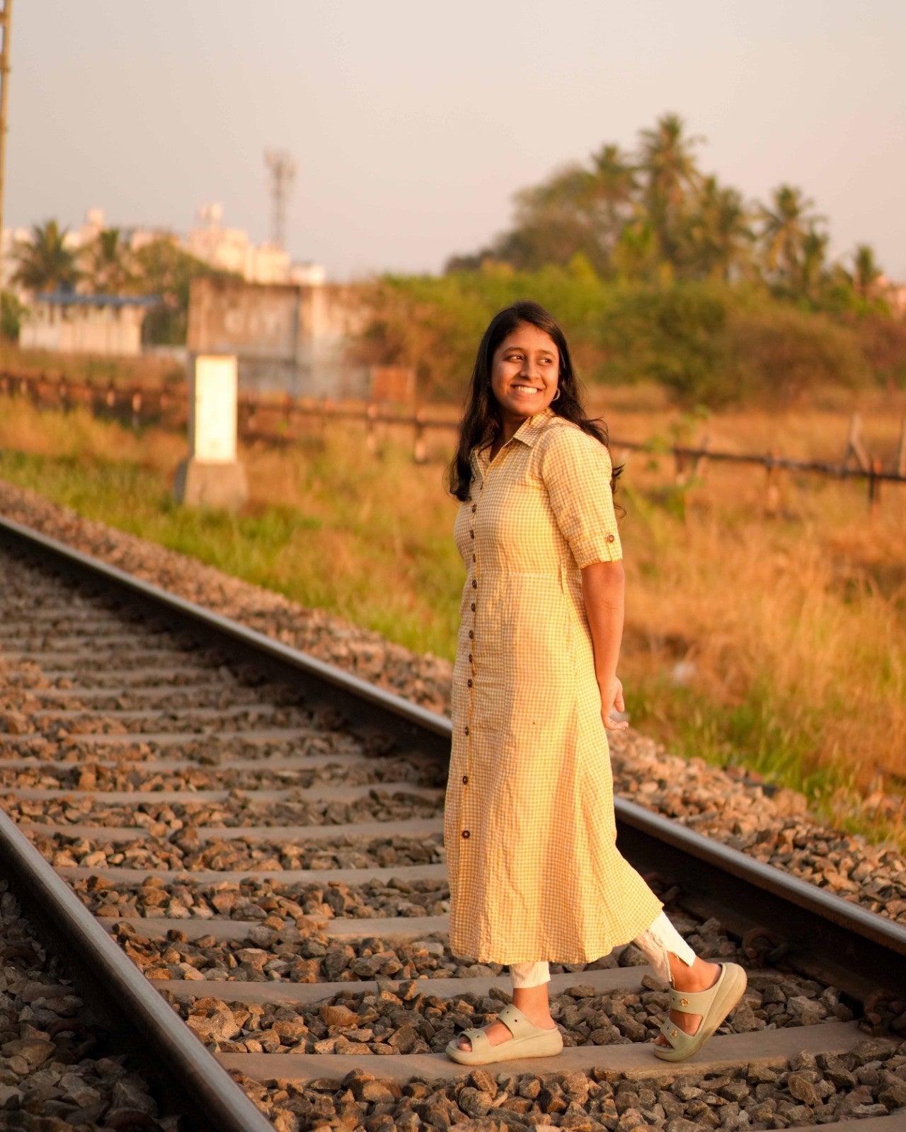 Side view of a woman in a yellow and white checked linen shirt dress, posing effortlessly on a railway track.